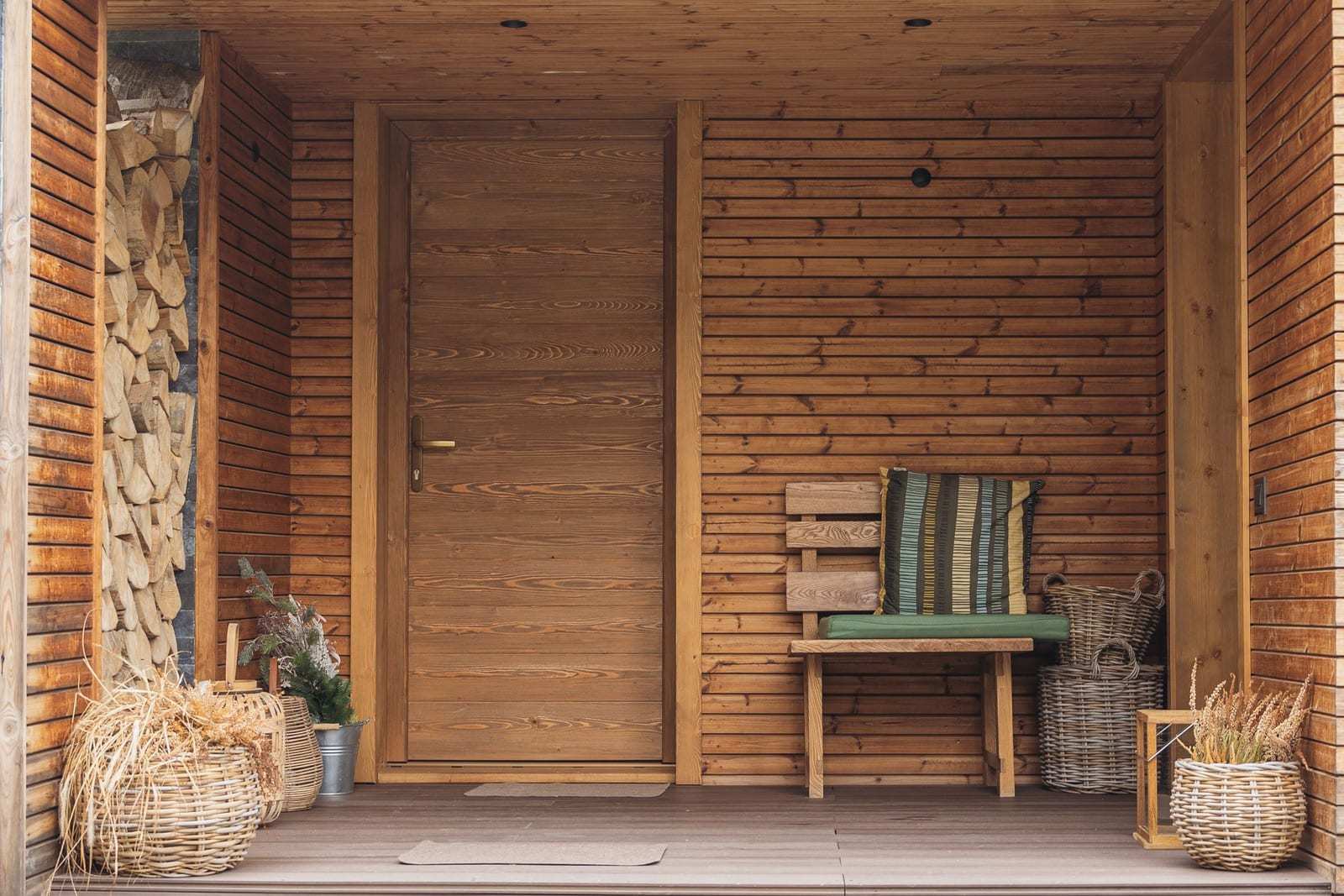 A covered cedar front porch with a wooden front door, a cushioned bench and woven baskets.