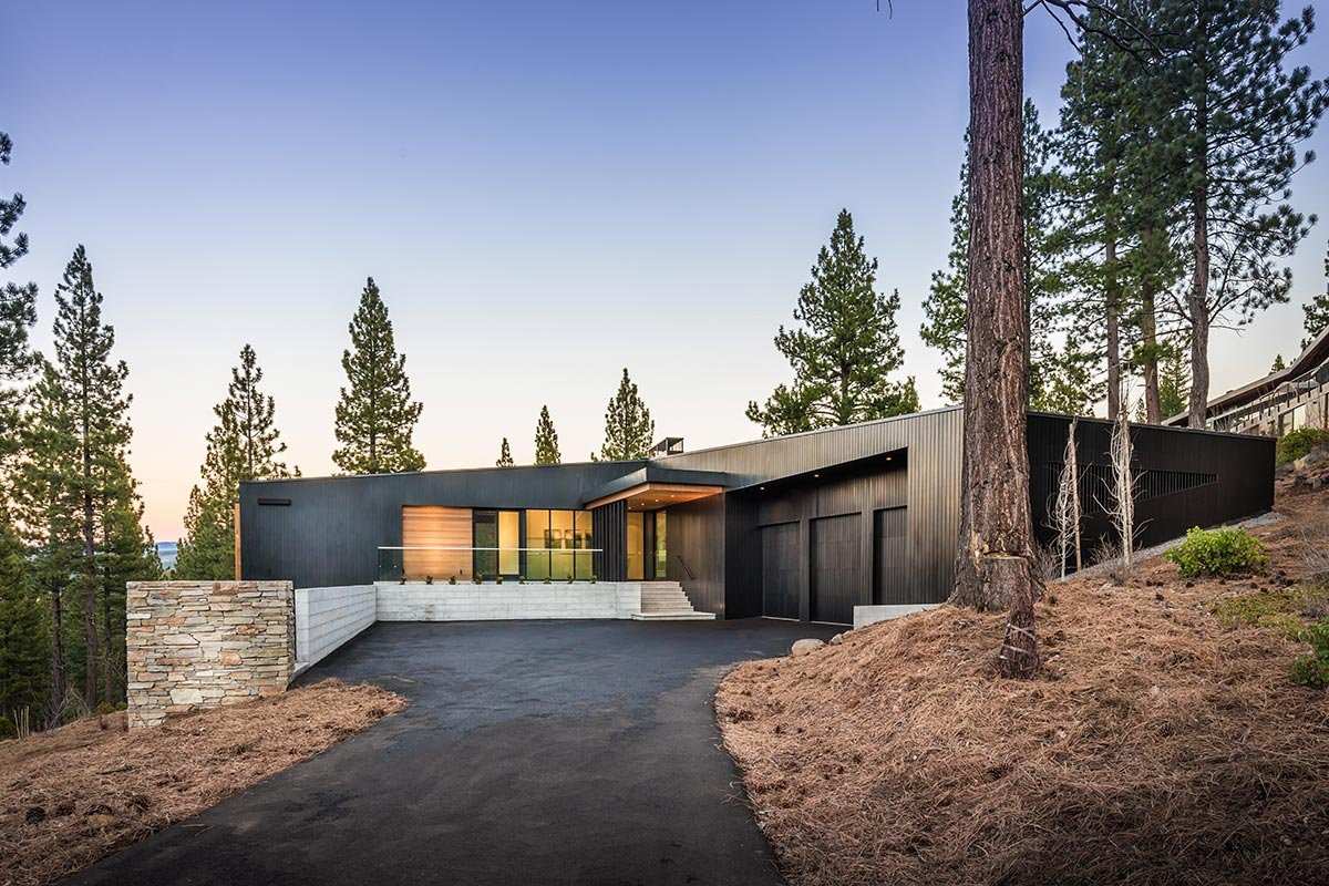 Modern mountain home with dark siding, stone wall and large windows among tall pines